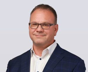 A man with short brown hair and glasses wearing a dark blue checked suit jacket and a white dress shirt, smiling slightly, poses against a light gray background.
