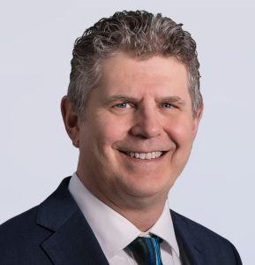 A middle-aged man with short, curly gray hair smiles at the camera. He is wearing a dark suit, a white shirt, and a blue patterned tie, against a plain light background.