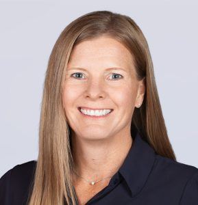 A woman with straight, light brown hair and blue eyes smiles at the camera. She is wearing a navy collared shirt and a delicate necklace, posed against a plain light grey background.