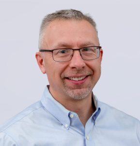 A middle-aged man with short gray hair and glasses is smiling. He is wearing a light blue button-up shirt and is posed in front of a plain, light-colored background.