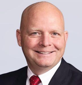 A smiling bald man in a dark suit, white shirt, and red patterned tie poses against a plain light gray background.