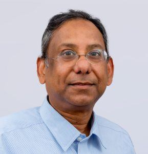 A middle-aged man with short dark hair, wearing glasses and a light blue collared shirt, looking at the camera and smiling slightly against a plain light background.
