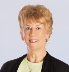 A smiling middle-aged woman with short, light brown hair, wearing a light green top, black jacket, and a necklace, posed against a plain light background.