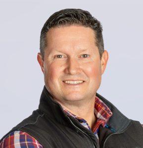 A man with short dark hair smiles at the camera. He is wearing a plaid shirt under a dark vest, and the background is light gray.