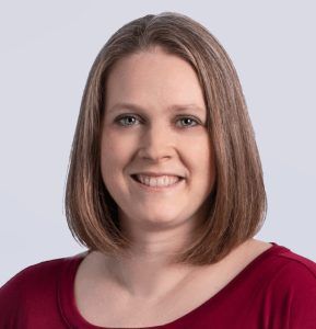A woman with straight, shoulder-length light brown hair smiles at the camera. She is wearing a red top and is posed in front of a plain light gray background.