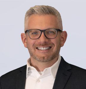 A smiling man with short gray hair and a trimmed beard wearing black glasses, a white collared shirt, and a dark suit jacket, against a plain light background.