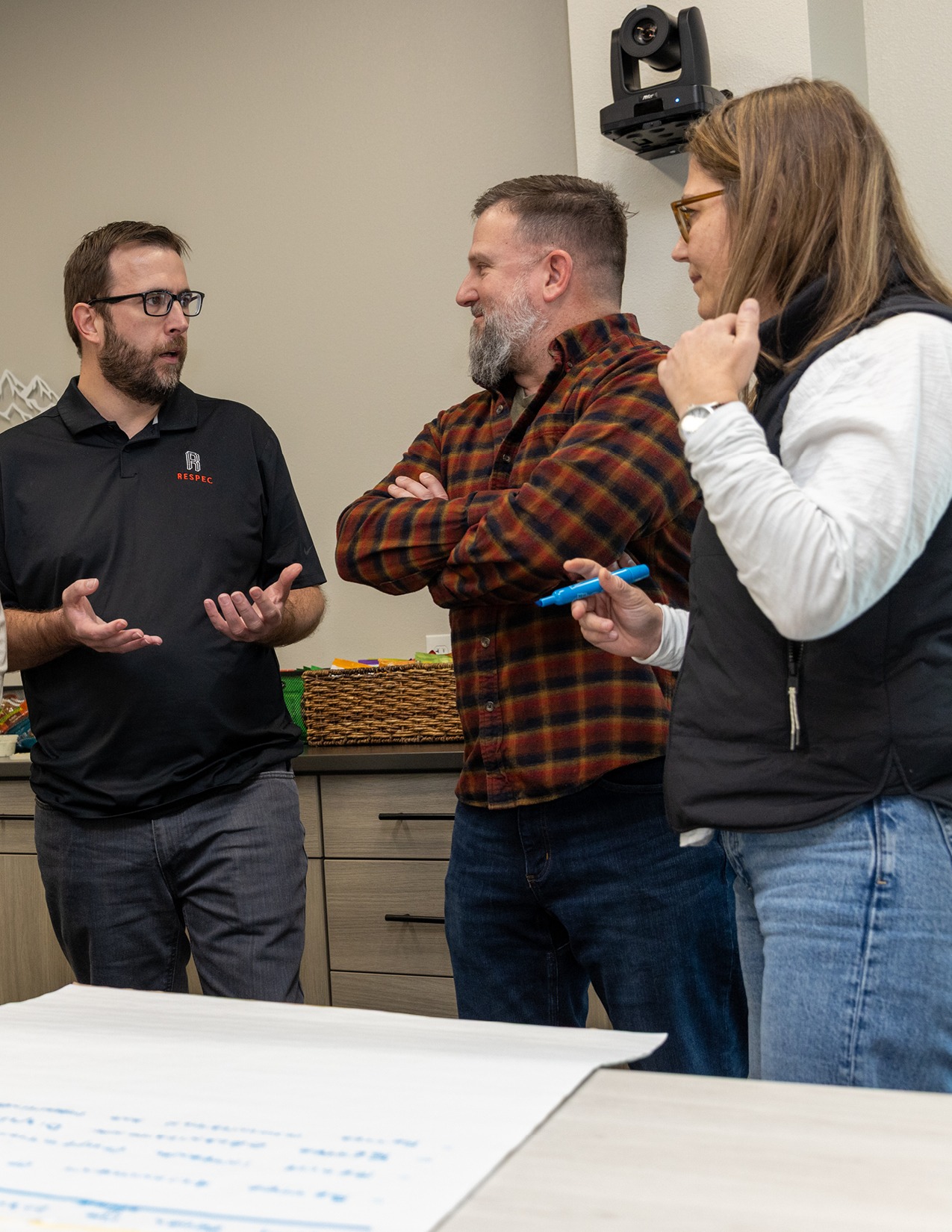 Three people stand and talk in an office-like setting near a table with a large sheet of paper covered in notes, actively engaged in discussion about engineering consulting and demonstrating thorough due diligence.