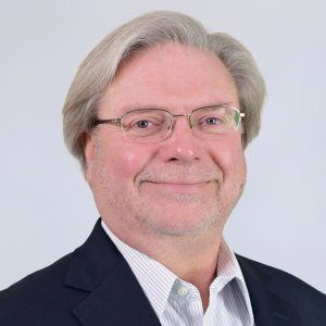 A middle-aged man with gray hair and glasses smiles at the camera. He is wearing a dark blazer over a light-colored collared shirt against a plain light background.