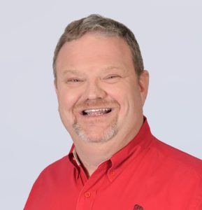 A middle-aged man with short light brown hair and a goatee, smiling widely. He is wearing a bright red collared shirt and is posed against a plain light gray background.