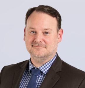 A man with short brown hair and a trimmed beard, wearing a brown suit, blue and white checkered shirt, and blue tie, smiles at the camera against a light gray background.