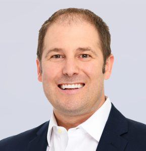Smiling man with short brown hair wearing a dark suit jacket and white collared shirt, against a light gray background.