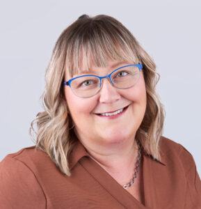 A smiling woman with shoulder-length blonde hair, wearing blue-framed glasses and a brown shirt, looks at the camera against a plain light gray background.