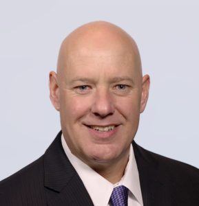 A bald man wearing a dark suit, white dress shirt, and purple tie, smiling at the camera against a plain light background.