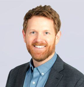 Smiling man with short red hair and beard, wearing a blue button-up shirt and a dark blazer, poses in front of a light gray background.