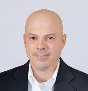 A man with a bald head, light skin, and brown eyes, wearing a white collared shirt and a dark blazer, is facing forward and smiling slightly in front of a plain light gray background.