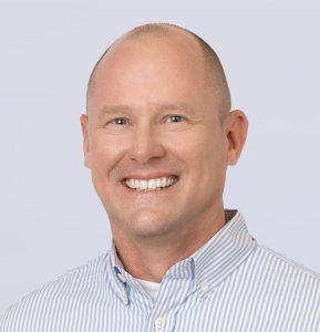 A smiling man with short light hair wears a blue and white striped button-up shirt, posing against a light gray background.