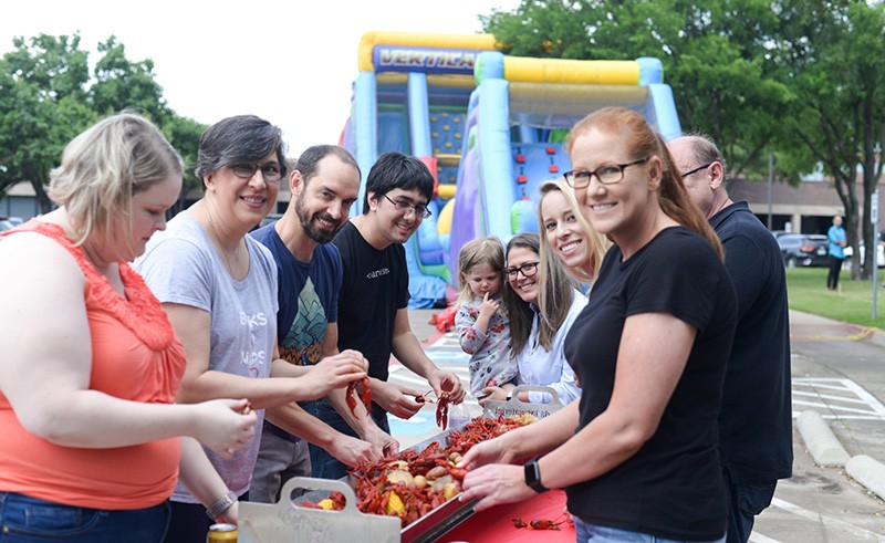 A group of people stand around a table outdoors, enjoying a crawfish boil. They are smiling and serving food. In the background, there is a colorful inflatable slide and green trees.