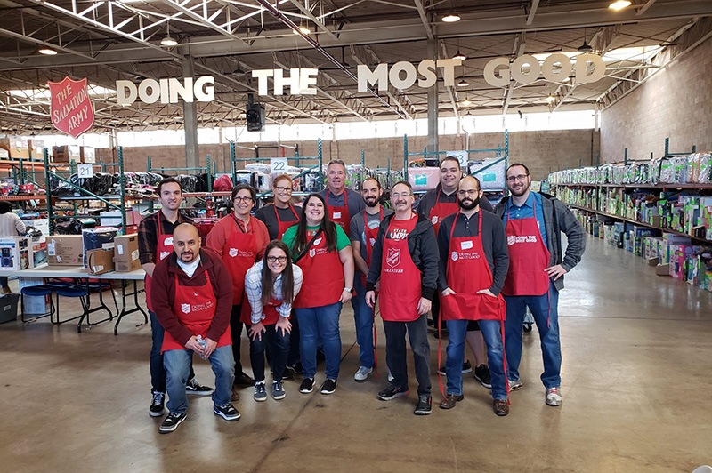 A group of people wearing red Salvation Army aprons pose and smile in a warehouse filled with toys. Large letters hang above them, reading “DOING THE MOST GOOD.”.