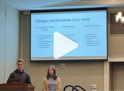 Two students stand at a podium giving a presentation. A projection screen behind them displays slides titled Changes and Decisions 2025-2026 with sections on Steel Bridge, Fundraising, and 2026 Symposium.