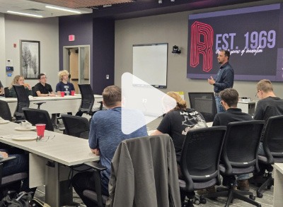 A man stands at the front of a conference room giving a presentation to a group of people seated at tables. A large screen behind him displays “EST. 1969” and “years of design.”.