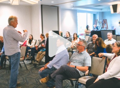 A man stands and speaks to a seated group of adults in a conference room, engaging them in discussion. The group appears attentive and relaxed, with natural light coming through a large window.