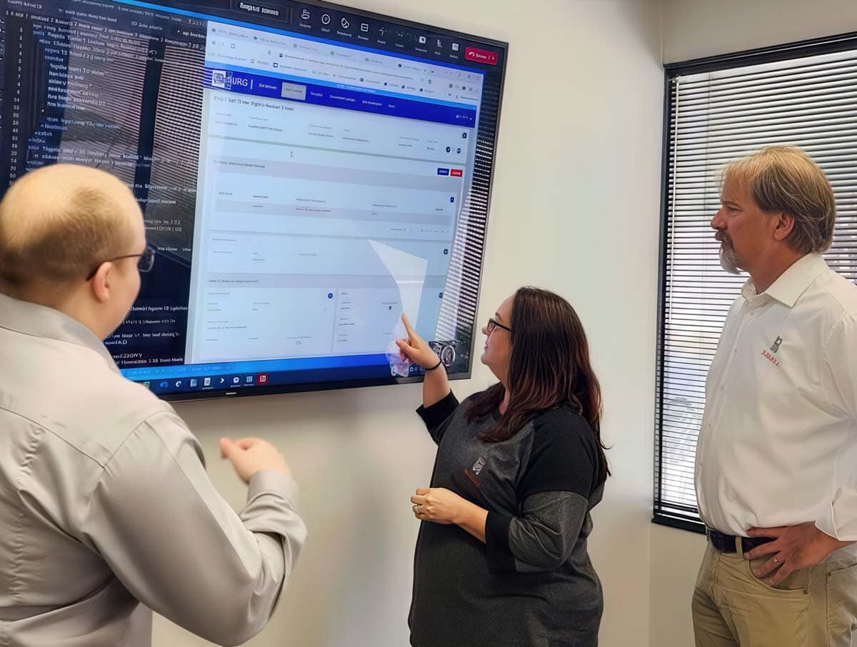 Three people stand in an office looking at a large wall-mounted screen displaying a web application. One woman points at the screen while the two men observe and discuss. Sunlight comes through window blinds.