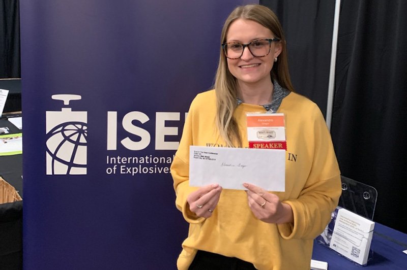 A woman with glasses and a yellow sweater holds an envelope and smiles. She stands in front of an ISEE (International Society of Explosives Engineers) banner, wearing event badges including one labeled SPEAKER.