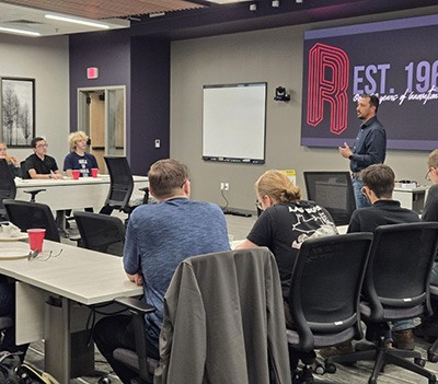 A man stands and speaks to a group of people seated at tables in a conference room. A large screen behind him displays EST. 1968 and part of a logo. The attendees listen attentively.