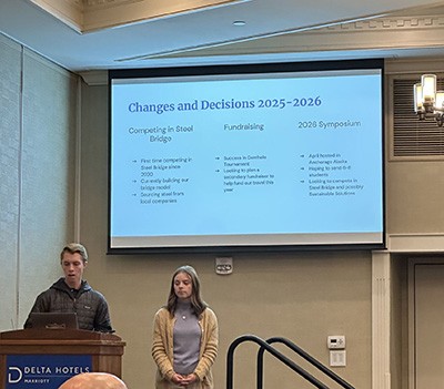 Two young adults stand at a podium presenting in a conference room. A projection screen behind them displays slides about “Changes and Decisions 2025–2026” covering competing in Steel Bridge, fundraising, and a 2028 symposium.