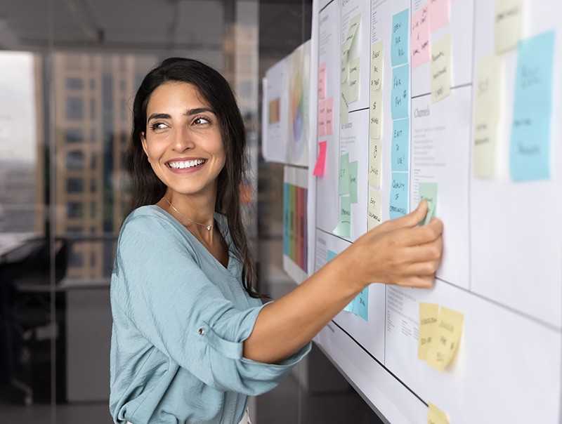 A woman smiles while placing sticky notes on a whiteboard covered with papers and colorful notes in an office setting. She appears engaged and happy, possibly brainstorming or planning a project.
