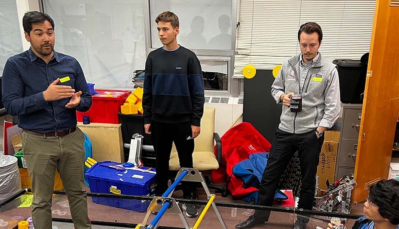 Three men stand in a cluttered room with boxes, chairs, and robotics equipment. One man gestures while speaking, another stands with hands at his sides, and the third leans against a desk holding a mug.