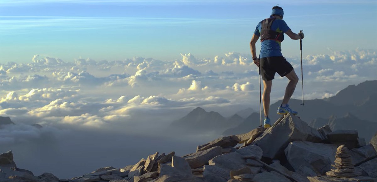 A person in athletic gear stands on rocky mountain terrain with trekking poles, overlooking a dramatic view of clouds and distant peaks under a blue sky at sunrise or sunset.