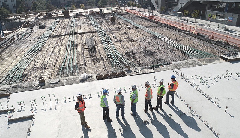 A group of six construction workers in safety vests and helmets stand together discussing on a large, partially built concrete foundation with exposed rebar and pipes at a construction site.