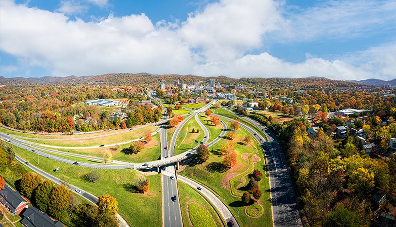 A panoramic aerial view of a city surrounded by rolling hills with autumn foliage, intersected by winding roads and highways under a partly cloudy sky. Cars travel along the roads, and scattered buildings dot the landscape.