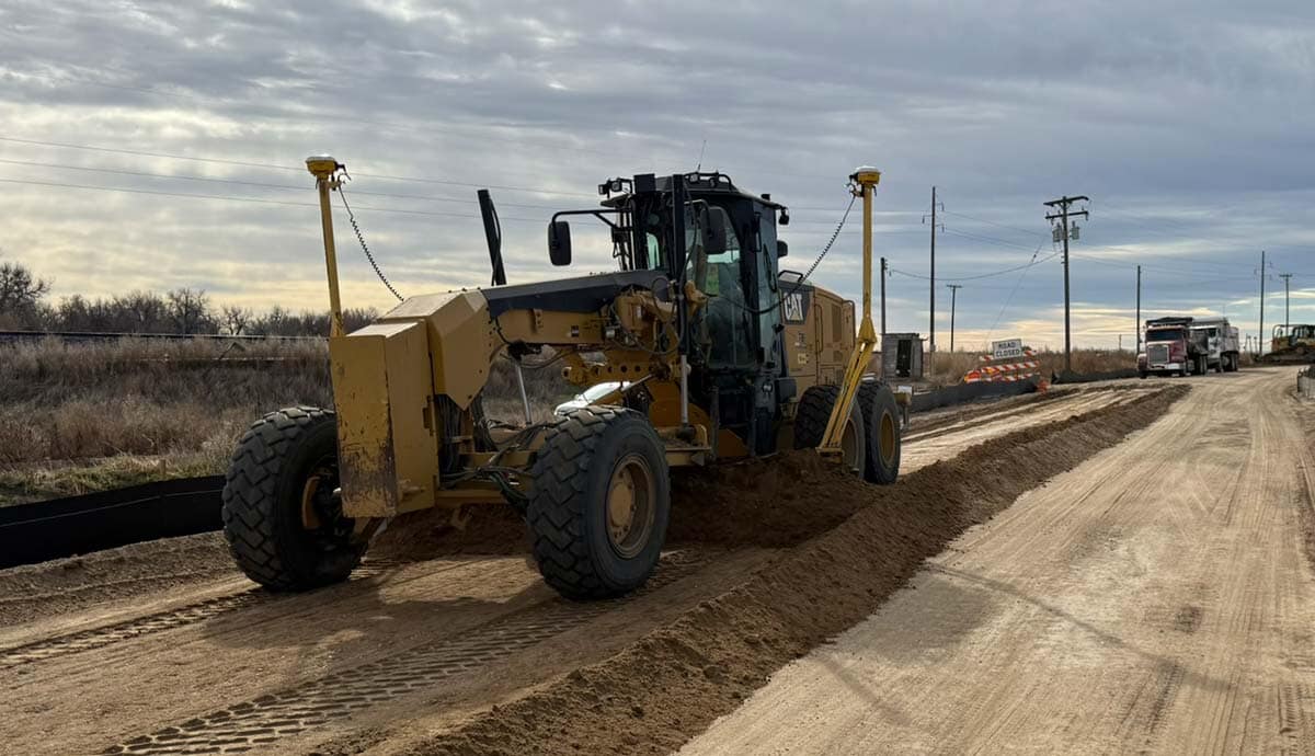 A yellow road grader smooths a dirt road under a cloudy sky, with construction barriers and a truck visible in the background. Utility poles and grassy fields line the roadside.