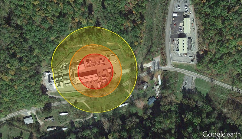 Aerial view of a facility surrounded by concentric red and yellow circles, indicating zones on the property. The facility is bordered by a forest, a road, and several buildings. Image is labeled Google Earth in the corner.