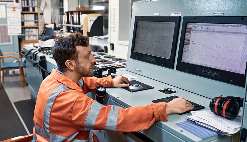 A man in an orange safety jacket sits at a control desk, monitoring data on two large computer screens in an industrial or control room setting. Documents and equipment are on the desk.