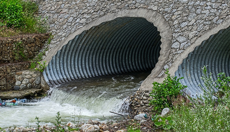 Two large corrugated metal pipes embedded in a stone embankment channel water through a culvert, with water flowing out from one pipe and surrounding green vegetation.