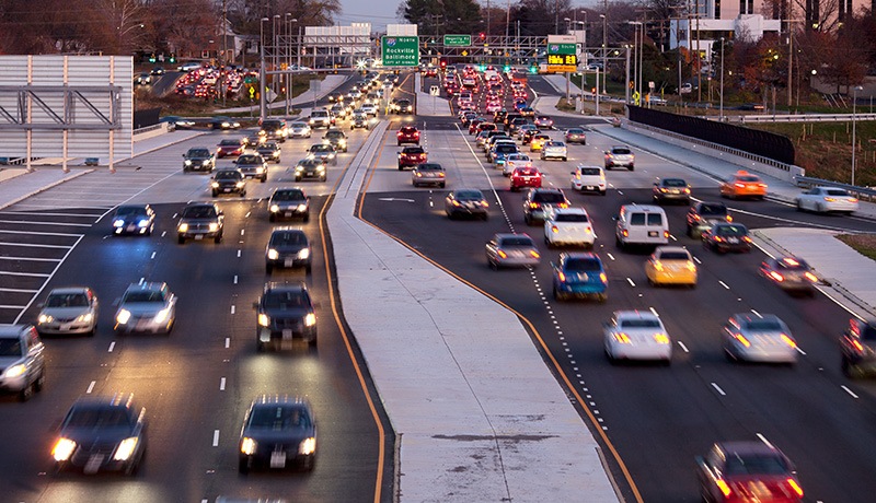 A multi-lane highway with heavy traffic in both directions during dusk, with cars headlights and taillights visible. Overhead signs and trees line the road, and buildings are seen in the background.