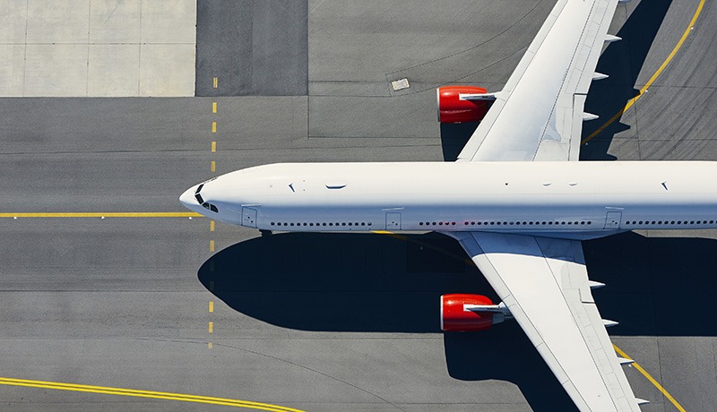 Aerial view of a white commercial airplane with red engines taxiing on an airport runway, surrounded by gray tarmac and yellow guiding lines.