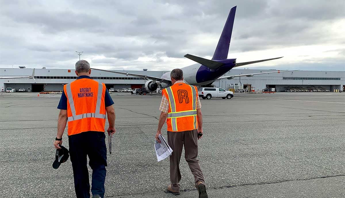Two airport workers in orange safety vests walk toward a large airplane on the tarmac under a cloudy sky, with airport buildings and vehicles in the background.