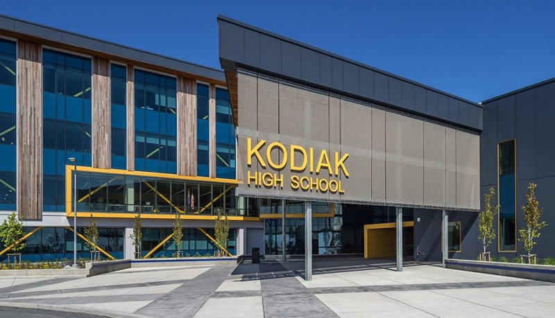 Modern school building with large windows and a prominent sign reading Kodiak High School above the main entrance. The exterior features yellow accents, and several young trees are planted near the entrance.