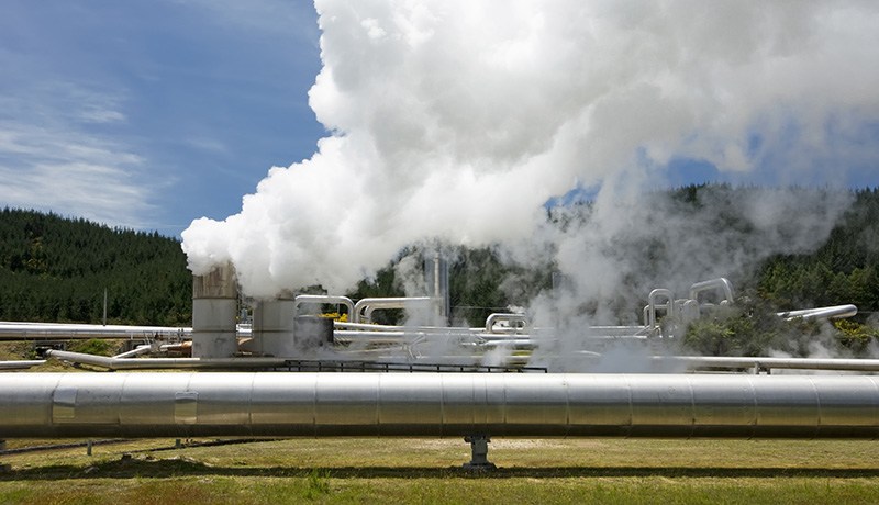 A geothermal power plant with large metal pipes and towers emitting white steam into the air, set against a backdrop of green trees and blue sky.