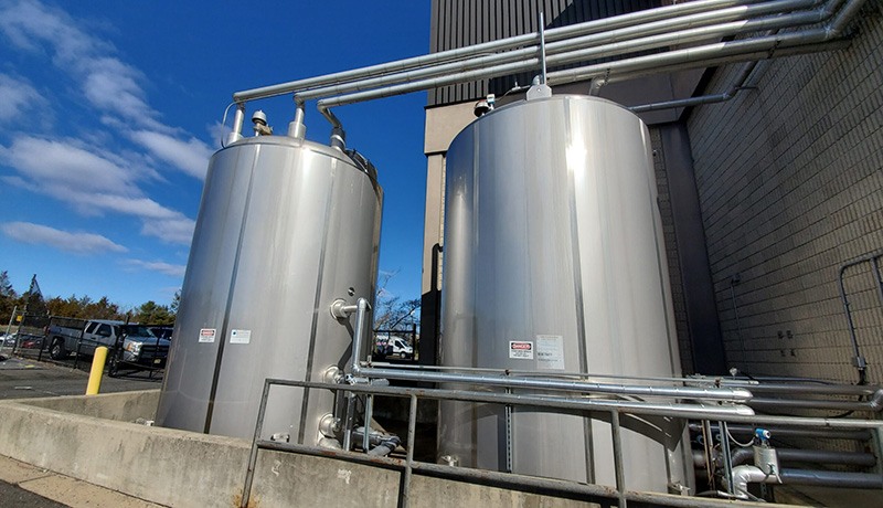 Two large, shiny stainless steel tanks with pipes attached, situated outdoors next to a brick building on a sunny day with a blue sky and scattered clouds.