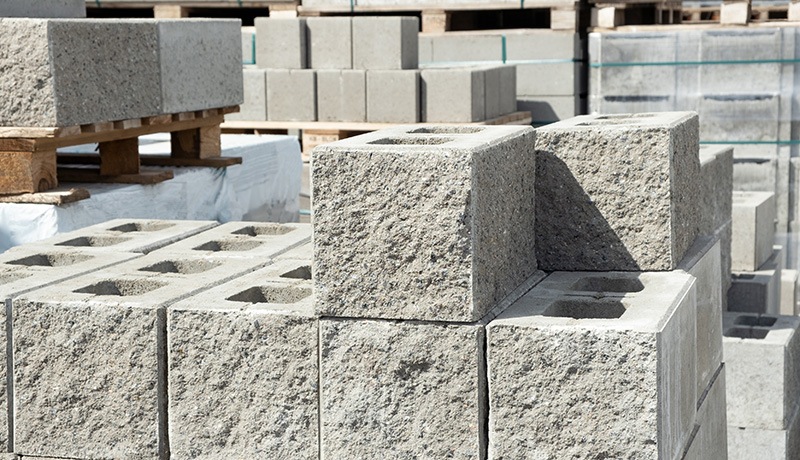 Several stacks of gray concrete cinder blocks are arranged outdoors on pallets at a construction supply yard. Some blocks have rough surfaces, and others have hollow centers.