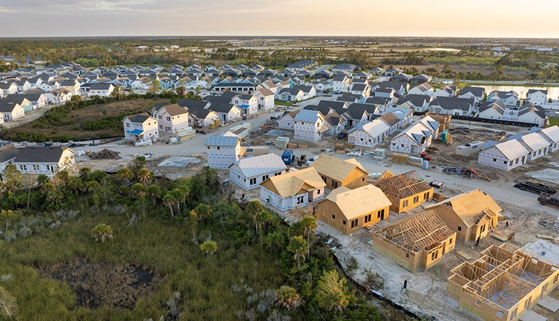 Aerial view of a suburban neighborhood with rows of completed houses and several homes under construction surrounded by green trees and open land at sunset.