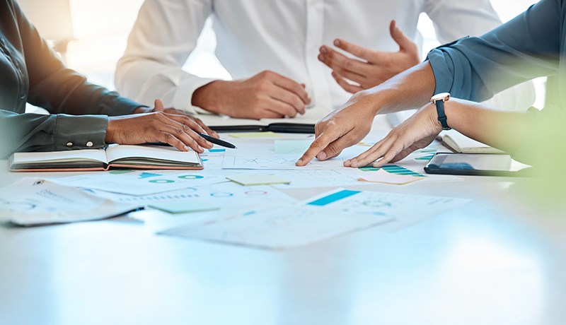 Three people sit at a table in a meeting, discussing documents and charts. One person gestures while two others point at papers with graphs and notes, indicating collaboration and planning.
