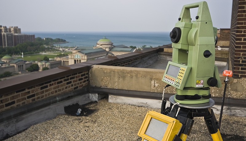 A green total station surveying instrument stands on a tripod on a rooftop, with a view of buildings, trees, and a body of water in the background under a clear sky.