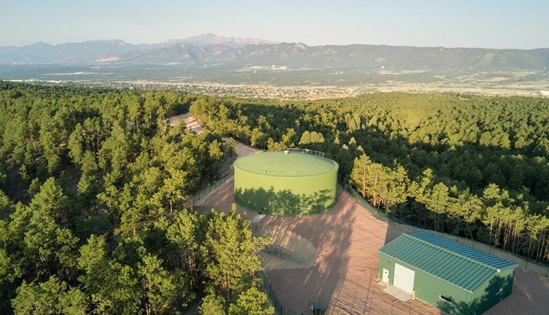 A large green water tank and a small green building sit among dense trees with mountains and a distant town visible in the background under a clear sky.