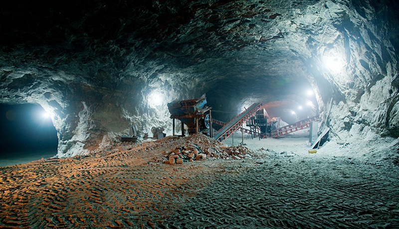 An underground mining tunnel with rough rocky walls, bright overhead lights, industrial conveyor belts, scattered machinery, and tire tracks on the dusty ground.